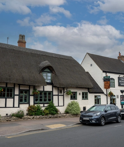 A view from across the road of the exterior facade and signage of The Royal Oak in Bishops Cleeve, with a thatched roof, and flower baskets hanging on the walls.