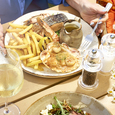 A banner showing a close up image of a person sat at a table with a seafood dish and glass of white wine within the exterior beer garden seating area to advertise the new menu at Chef & Brewer venues.