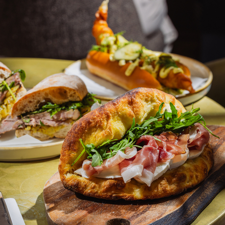An advertising image showing 2 plated sandwich dishes and a sandwich dish presented on a wooden board sat on a table within the interior restaurant seating area at The Four Oaks.
