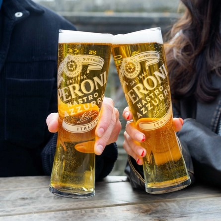 Two people sitting at a table in a beer garden, holding glasses of Peroni beer.