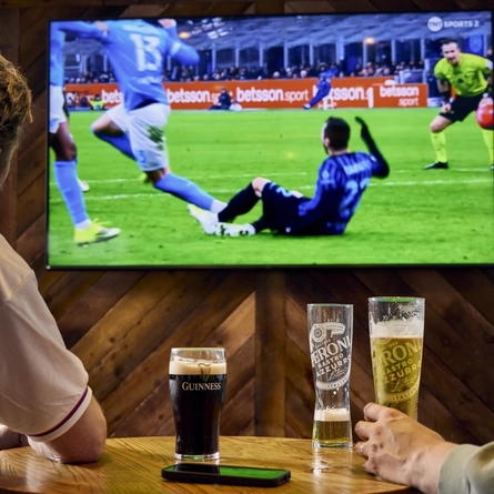 Two people sitting at a table in a pub, watching sports on the TV on the wall in front of them. A glass of Guinness, two glasses of Peroni, and a mobile phone sit on the table.
