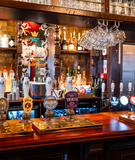 A close up view of the beer taps and golden drinks trays on the bar inside the Two Chairmen in St James's, with glass tankards hanging above the counter, and bottles of wine and alcoholic spirits on shelves behind the bar.