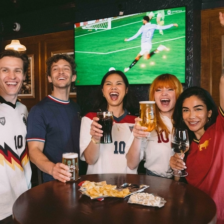 An image showing a group of friends stood at a table watching a football match and enjoying a selection of drinks and snacks while celebrating within the interior seating area at a Greene King venue.