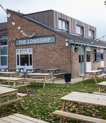 The exterior facade, signage, and beer garden seating area at The Longship in Hebburn, with wooden picnic tables, string lights overhead, and a TV on the wall.