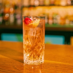 A close up view of an Another Raspberry Collins cocktail served in a highball glass with ice, dried raspberries and a lemon wedge. The glass stands on the bar inside a pub.