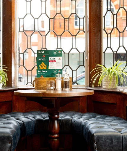A close up view of a circular restaurant table with an upholstered U-shaped booth seat in a small leaded, bay window at The Talbot. Menus, salt and pepper mills, and a tealight stand on the table; and two potted plants sit on the windowsill.