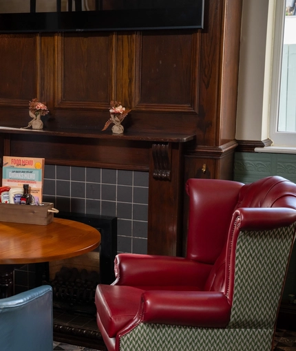 A close up view of a small, round restaurant table next to a dark wood fireplace inside The Longship, with two large, wingback chairs, and a TV on the wall above the fireplace.