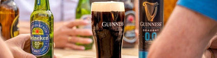 A wooden table with a can and pint of Guinness 0.0 Draught in the centre. Surrounding the table are the blurred out torsos of four people, each holding different drinks. 