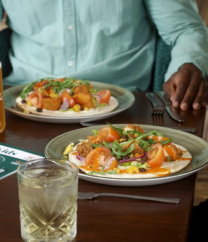 A table with two plates of flatbreads topped with vegetables and crispy pieces, accompanied by drinks including beer and a cocktail. Two people seated at the table have their hands visible near the food.