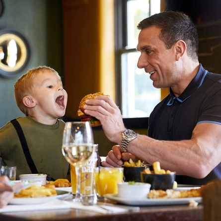 A man seated at an indoor table, holding a burger to a child.