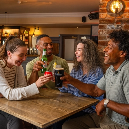 Four people gathered around a wooden table in a pub, raising assorted drinks in a toast. The drinks include a pint of Guinness, a bottled alcohol-free beer, a can of soft drink and a mixed drink with ice and lemon. The pub interior features exposed brickwork, a stocked bar, warm lighting and wooden furnishings.