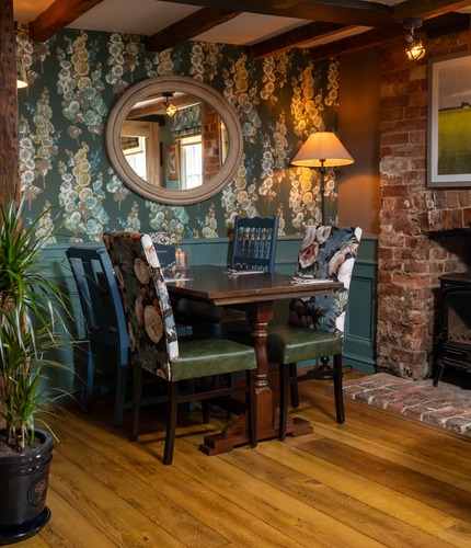 An interior restaurant seating area at The Kings Arms in Sandford-On-Thames, with wooden flooring and beams, upholstered chairs, and a brick fireplace with a log burner.