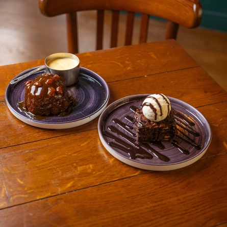 A Sticky Toffee Pudding served on a plate with a pot of custard, and a Brownie served on a plate with a scoop of ice cream and drizzled with sauce. The plates sit on a wooden restaurant table.