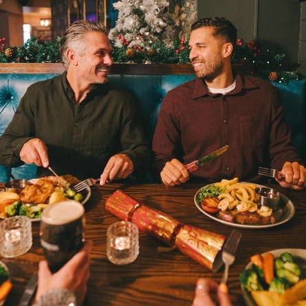 Four people sitting at a restaurant table with plates of food, Christmas crackers, a glass of red wine, and a glass of champagne in front of them. One person is holding a glass of stout, and Christmas decorations are visible in the background.