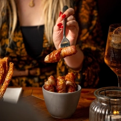A person is holding a fork with a piece of Pigs in Blankets over a small gray bowl. 