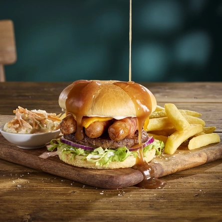 The Gravy Pig Dipper burger served on a wooden serving board with fries and coleslaw. The board sits on a wooden table, and gravy is being poured over the burger.