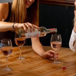 A woman is seated at an indoor table, pouring a glass of rose wine from a bottle into her companion's glass. 