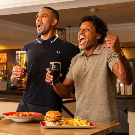Two people standing beside a high table in a pub, holding a pint of lager and a pint of Guinness. Plates of food, including a burger with chips and a pasta dish, are on the table. The pub interior features a stocked bar, exposed brickwork and warm lighting in the background.