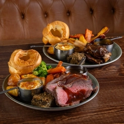 Two plates of roast dinner, including meat, Yorkshire pudding, vegetables and gravy, sitting on a wooden table.