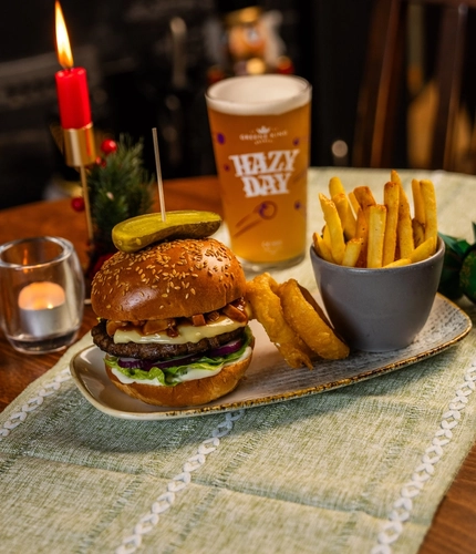 A Festive Burger served on a plate with onion rings and a bowl of fries. The plate sits on a wooden restaurant table along with a glass of Hazy Day, a Christmas cracker, a tealight, a lit red taper candle, and a small glass vase of dried flowers; and a Christmas nutcracker is visible in the background.