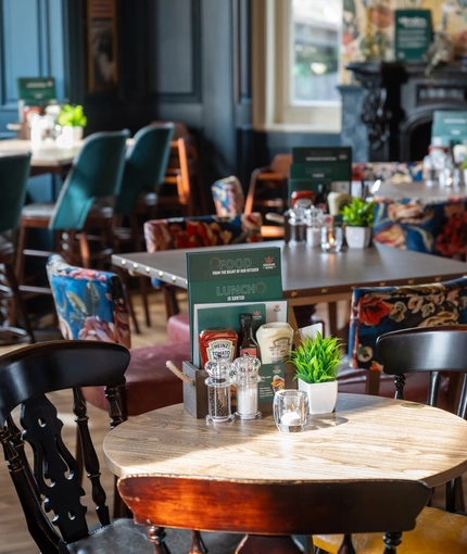 Menus, condiments, a tealight, and a potted plant, on a restaurant table inside The Victoria. A fireplace and other restaurant tables are visible in the background.
