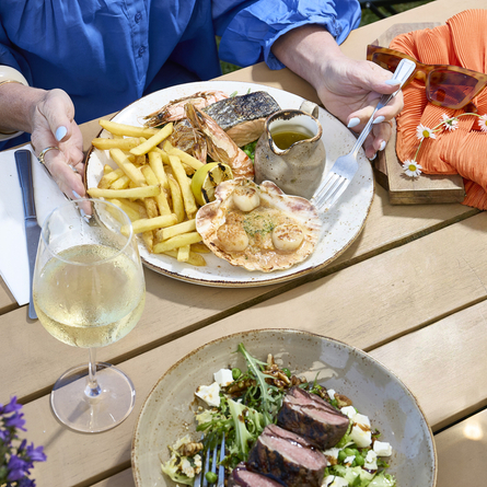 A lifestyle image showing a person sat at a table enjoying a Seafood Platter dish and a glass of white wine within the exterior beer garden seating area at a Chef & Brewer venue.