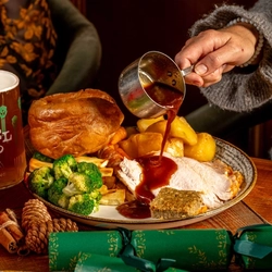 A wooden table holding a plated roast turkey dinner.  A hand is holding holding a small pan of gravy pouring of the plate.