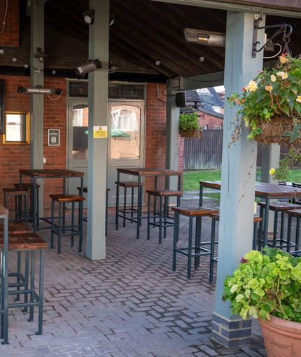 An exterior under cover beer garden seating area at the Green Posts, with a TV on the wall, string lights above the tables, and hanging flower baskets on the wooden support beams.