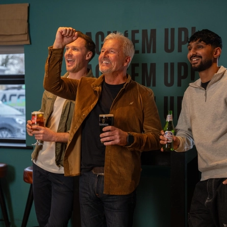 Three people standing in the gaming area of a restaurant. One of them is holding a glass of beer, one is holding a glass of Guinness, and the other is holding a bottle of Peroni.
