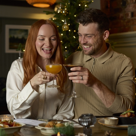 A banner showing 2 people holding cocktails and celebrating with a cheers with plated picky bits sat on the table to advertise Christmas at Chef & Brewer.