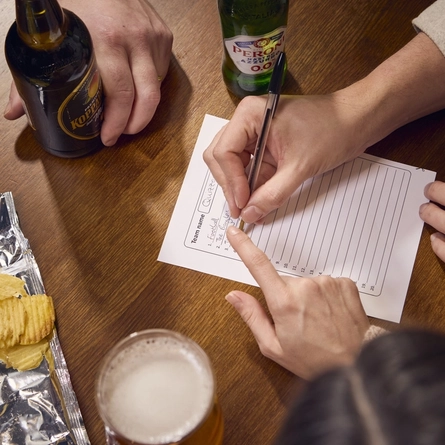 A bird's eye view of people at a table filling out a pub quiz question sheet. Bottles of Peroni 0.0% alcohol beer and Koppaberg cider, a glass of beer, and an open packet of crisps also sit on the table.