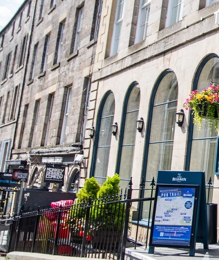 The exterior facade, signage and seating area of The Advocate in Edinburgh, with hanging flower baskets on either side of the entrance.