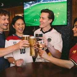 An image showing a group of friends stood at a table watching a football match and enjoying a selection of drinks while celebrating within the interior seating area at a Greene King venue.