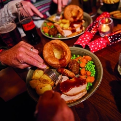 A festive dinner table with plates of food, including roasted vegetables, Yorkshire pudding, and slices of meat. The table is decorated with red napkins, Christmas crackers, and a lit candle. There are drinks on the table including a bottle of Sprite and a glass of water with lemon. Several hands are seen holding utensils and eating from the plates