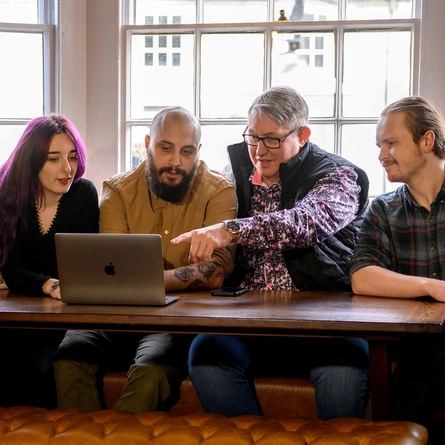 Three Men and a Woman sat at a table looking at the laptop in front of them, The Man in the middle is pointing towards the laptop