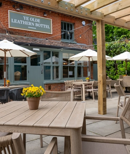 The paved exterior beer garden seating area at the Ye Olde Leathern Bottel in Barkham, with an under cover section, shade umbrellas, potted plants on each table, and string lights overhead.