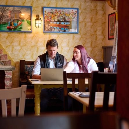An image showing 2 people sat at a table within the interior seating area looking at a laptop and talking for Pub Partners.