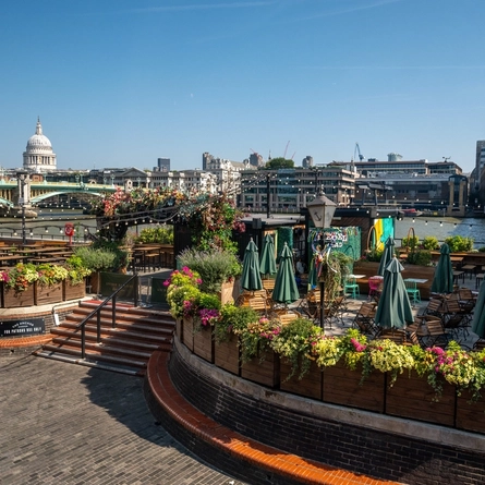 The exterior beer garden seating area at The Anchor Bankside in Southwark; with shade umbrellas on some of the tables; string lights hanging overhead; a view of the River Thames; and steps leading up to the seating area which is surrounded by planter boxes filled with flowers. 