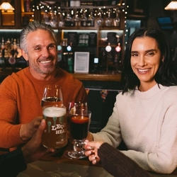 Four people sitting at a table in a bar, clinking together glasses of white wine, mulled wine, Level Head, and a cocktail. A Christmas tree decorated in red and gold stands beside the table, and fairy lights are visible in the background.