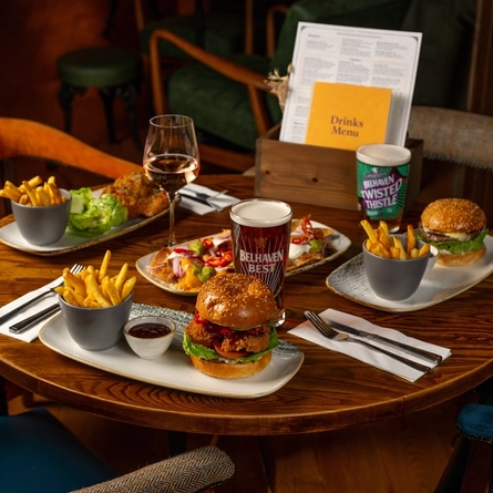 A round, wooden restaurant table laden with plates of food including burgers, fries, nachos, and salad. A glass of wine, a glass of Belhaven Best ale, a glass of Belhaven Twisted Thistle IPA, cutlery, and menus also sit on the table.