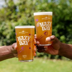 Two hands, each holding a pint of Hazy Day in the air, against a backdrop of greenery in a beer garden.