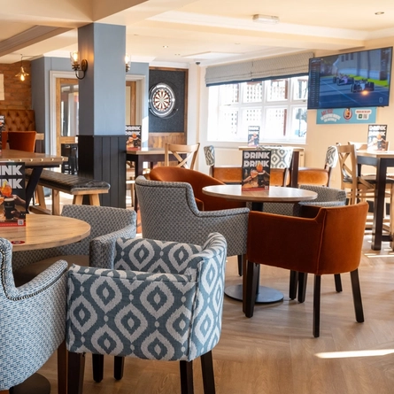The interior restaurant seating area at The Weathervane in Stoke On Trent, with upholstered chairs and booth seats, a dartboard, and a TV on the wall.