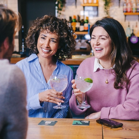 A group of friends sat in the interior seating area of a Greene King pub enjoying glasses of Gin at the Gin weekend takeover event. 