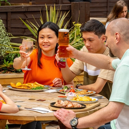 A family seated at an outdoor table in a beer garden, holding their drinks in a Cheers pose whilst enjoying a meal..
