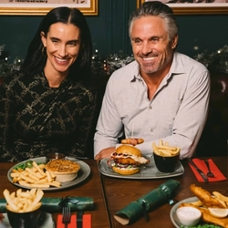 Two people sitting at a restaurant table with a plate of pie, chips, peas, and gravy; and a plate of a burger and chips in front of them. Christmas crackers, other plates of food, and glasses of Level Head, Hazy Day, and white wine also sit on the table; and a Christmas garland lit with fairy lights is visible in the background.