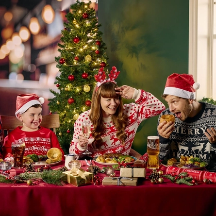 Three adults and a child sitting side by side at a restaurant table and eating from the plates of Christmas dinner in front of them. Glasses of beer and fizzy drink stand on the table along with Christmas presents, Christmas crackers, baubles, and pine cones; two of the adults are holding glasses of wine; and a Christmas tree is visible in the background.