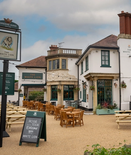 The exterior facade, signage, and beer garden seating area of The White Lion in Frenchay, with wooden picnic tables, and hanging flower baskets on the front of the building. 