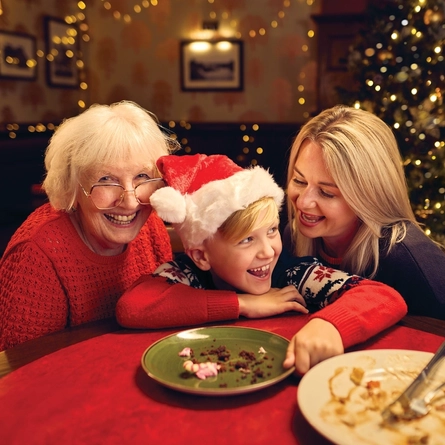 A child and two women sitting at a table inside a pub, empty plates and cutlery sitting on the table in front of them. A Christmas tree and fairy lights are visible in the background.