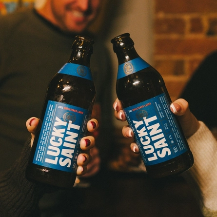 Two friends holding a bottle of low and no Lucky Saint beers in a cheers in a Greene King venue.