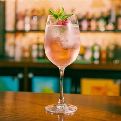 A close up view of a Rosé Hugo Spritz cocktail served in a wine glass with ice, mint, and dried raspberries. The glass stands on the bar inside a pub.